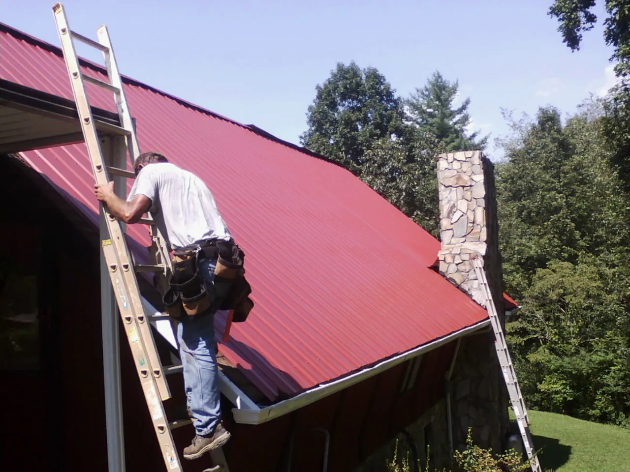 Roofer on a ladder beside a red metal roof and stone chimney — Soffit & Fascia Repair by Roofing Brothers in Alberton
